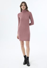 Strick-Turtleneck-Kleid in Hellrosa mit Zopfmuster. Figurbetonte Form, bis zur Mitte des Oberschenkels, kombiniert mit schwarzen Stiefeletten.