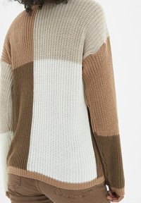 Pull en maille aux blocs de couleurs marron, beige et blanc. Présente une coupe ample avec un tissu texturé et des poignets et un ourlet inférieur côtelés.