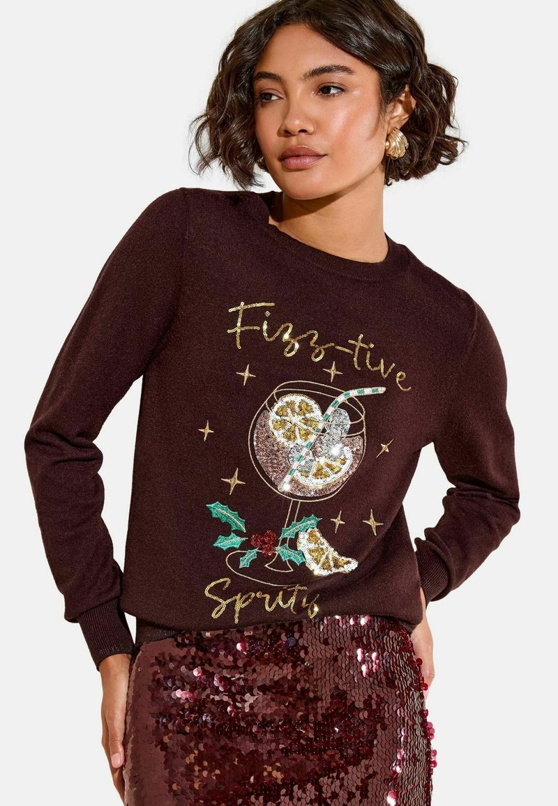 Maglione bordeaux con una grafica di una bevanda festiva decorata con paillettes, lettering dorato e decorazioni di agrifoglio, abbinato a una gonna di paillettes scintillante.