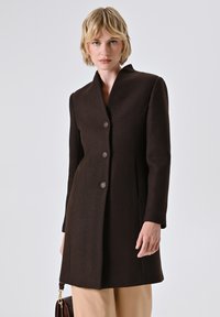 Vrouw met kort blond haar, gekleed in een donkerbruine knopenjas en beige broek, met een bruine handtas tegen een effen achtergrond.