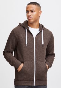 Brun lynlås hoodie lavet af tekstureret stof, med en hvid lynlås, hvide snore og forlommer. Med hætte.