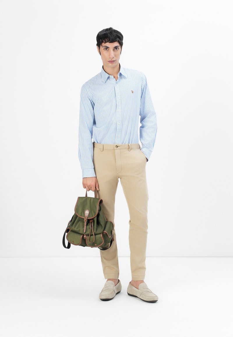 Jasnoniebieska koszula w paski z guzikami, beżowe spodnie, beżowe mokasyny, trzymająca zielony plecak z brązowymi akcentami. Prosty design, casualowy styl.