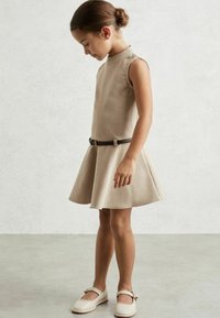 Vestido sin mangas beige con un cuerpo ajustado y falda acampanada, ceñido a la cintura con un cinturón marrón oscuro. Zapatos claros con lazos.