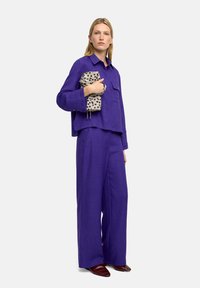 Fioletowa lniana oversize bluzka w komplecie z szerokimi spodniami. Modelka trzyma kremową torebkę w czarne kropki. Ciemnoczerwone płaskie buty.
