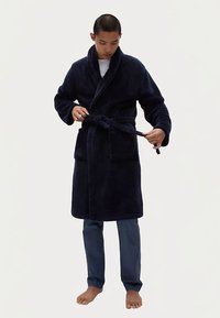 Námořnicky modrý plyšový župan s klopovým límcem, pásem v pase a dvěma předními kapsami. Model stojí bosý a má na sobě šedé kalhoty.