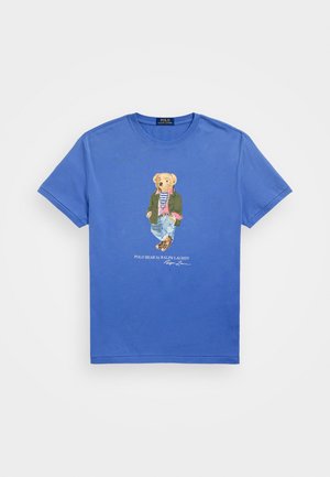 Zila kokvilnas T-krekls ar detalizētu grafiku, kurā iekļauts lācis neformālā apģērbā, ar tekstu "POLO BEAR by RALPH LAUREN" zem tā.