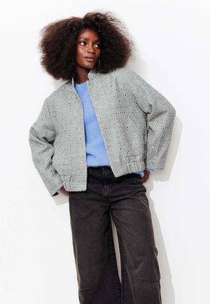 Veste bomber grise texturée avec une coupe ample, col montant et accents métalliques subtils, associée à un pull bleu et un jean foncé.