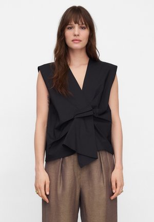 Femme aux cheveux longs et bruns portant un haut noir sans manches à col châle large et un pantalon brun ample, debout devant un fond blanc uni.
