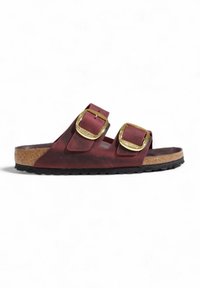 Donkerrode slide-sandalen met twee grote gouden gespen, kurken middenzool en zwarte gehanteerde zool op een witte achtergrond.