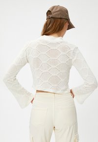Eine Frau trägt einen weißen, hexagonalen, gestrickten Cropped-Top, beige, hochtaillierte Hose und eine braune, karierte Mütze, von hinten gesehen.