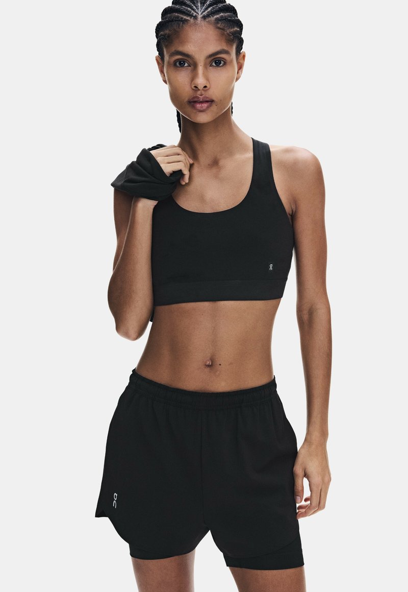 Femme aux cheveux tressés portant un soutien-gorge de sport noir et un short, tenant un vêtement noir sur son épaule.