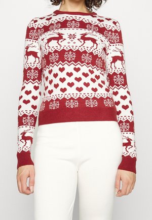 Rode en witte trui met rendieren, sneeuwvlokken en hartpatronen, gedragen met een witte broek, zichtbaar van schouders tot heupen.