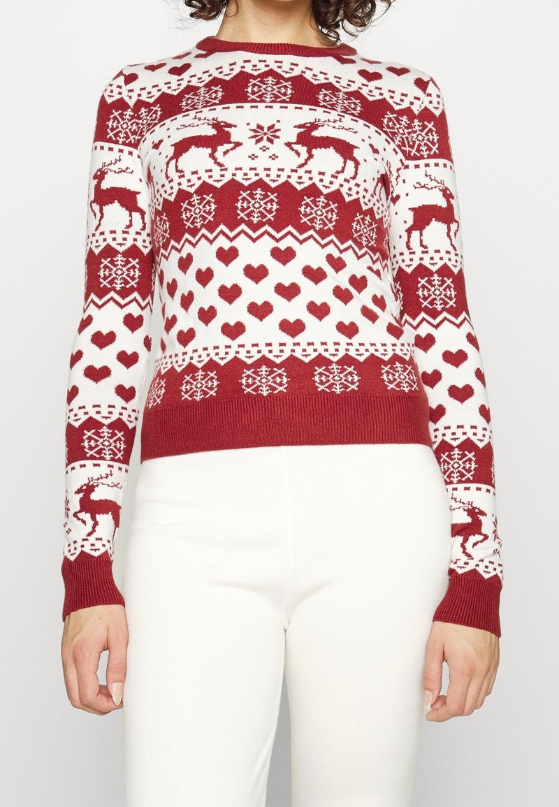 Pull rouge et blanc avec des motifs de rennes, flocons de neige et cœurs, porté avec un pantalon blanc, visible des épaules jusqu'aux hanches.
