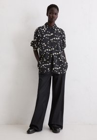 Zwarte bloemenoverhemd met witte accenten, knoopsluiting, ontspannen pasvorm. Gecombineerd met wijde zwarte broeken en zwarte schoenen.