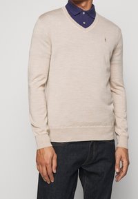 Beige v-ringad tröja i mjukt stickat material, med långa ärmar och en liten broderad logotyp på bröstet, ihop med en marinblå skjorta med krage.