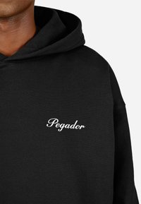 Felpa nera con cappuccio realizzata in tessuto morbido, dotata di una tasca a marsupio sul davanti e un logo "Pegador" bianco nella zona sinistra del petto.