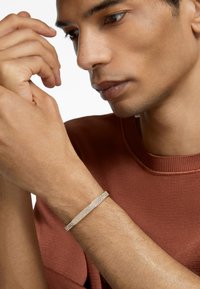 Bracciale in metallo dorato con superficie texturizzata e gemme trasparenti incastonate, indossato su un polso, in contrasto con un maglione color ruggine.