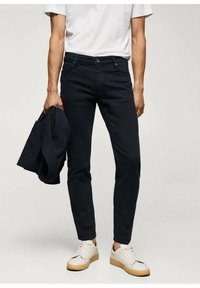 Tmavě modré slim-fit džíny s pěti kapsami, v kombinaci s bílým tričkem a světlými teniskami. V levé ruce drží odpovídající bundu.