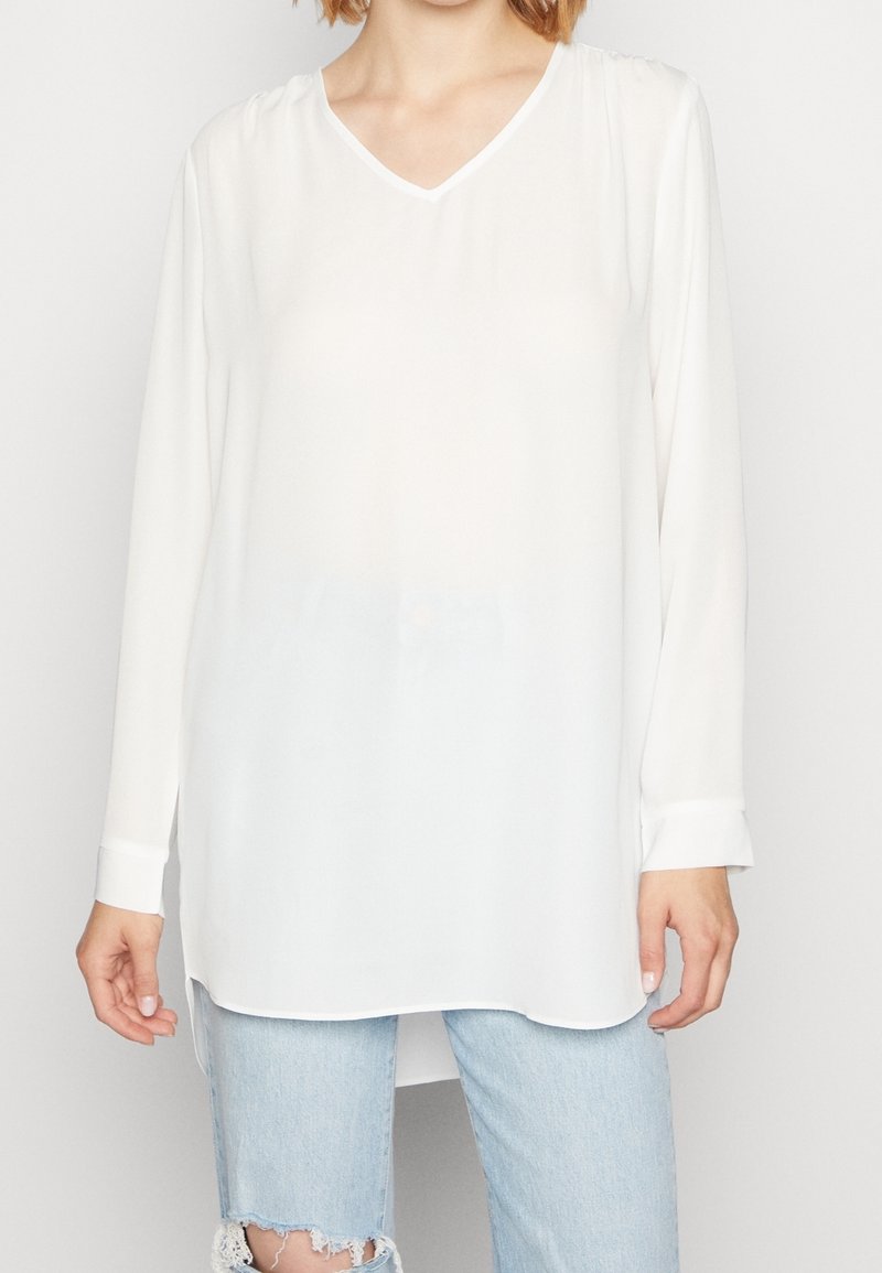 Femme portant un chemisier blanc ample à manches longues avec un col en V et un jean bleu clair déchiré, montrée du cou aux cuisses.