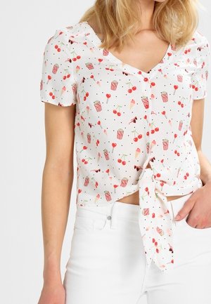 Biała bluzka z krótkim rękawem, z wiązaniem z przodu, zdobiona wzorami czerwonych wiśni, rożków lodów, popcornu i lizaków, noszona z białymi spodniami.