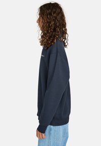 Marineblauwe sweatshirt met een ronde hals en verlaagde schouders. Bevat ribgebreide boorden en onderrand. Draagt in combinatie met lichtblauwe jeans met een textuur.