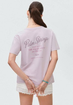 T-shirt en coton violet clair avec un motif de texte mélangé au dos, à manches courtes et coupe décontractée. Porté avec un short en dentelle blanche.