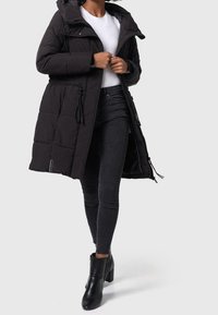 Sort polstret lang frakke med hætte, elastisk talje, lynlås lukning og snore. Kombineret med sorte stramme jeans og støvler med hæl.