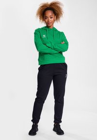 Grüne Kapuzenjacke mit Kordelzug und frontalem Taschen, kombiniert mit schwarzen, schmalen Jogginghosen mit kleinem Logo. Getragen mit schwarzen Sportschuhen.