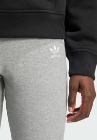 Sive rebraste tajice s bijelim adidas logotipom, u paru s crnom duksericom koja ima rebrasti završetak rukava. Ruka resting na području bedara.