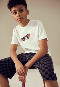 Hvid t-shirt med farverig skateboard-grafik, parret med sorte og grå ternede shorts. Fast materiale, afslappet pasform, korteærmer.