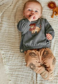 Smilende baby ligger på en cremefarvet tæppe, iført en grå bluse med løvemed grafisk motiv og teksten "Vær modig", brune bukser med løvemotiver og beige sko.