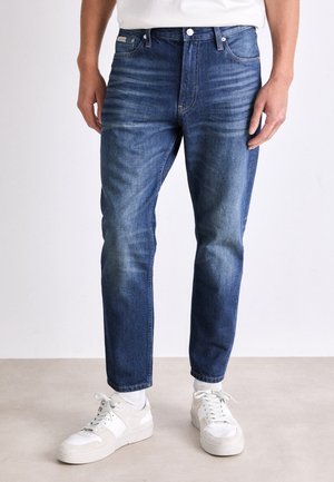Denim jeans i medium blå med subtil falmning, fem lommer og lige snit. Sammen med hvide sneakers og lyse sokker.