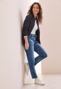 Tenue décontractée comprenant une veste bomber noire, un t-shirt blanc, un jean bleu clair et des baskets blanches avec des accents gris clair.
