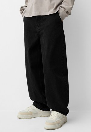 Person iført oversize sorte bukser og hvide sneakers med beige accenter, stående med hænderne i lommerne mod en hvid baggrund.