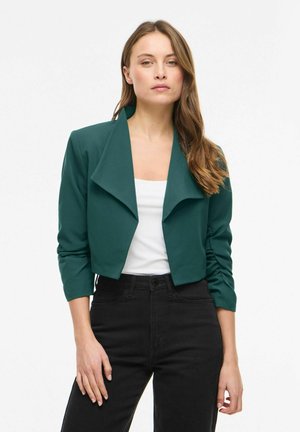 Kvinne som har på seg en kort mørkegrønn blazer over en hvit topp og høytlivede svarte bukser, stående foran en ensfarget hvit bakgrunn.