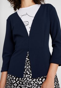 Navy long-sleeve cardigan with a V-neck, fitted design, and textured fabric, worn over a white shirt with printed accents and a leopard-print skirt.