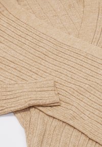 Beige bordás pulóver, V kivágással, textúrált mintával. Az anyaga puhanak és kötöttnek tűnik, enyhe rugalmassággal.
