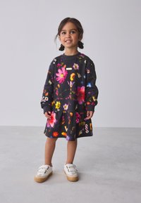 Mustast lillemustriga dressipluus, millel on erksad lillemustrid, pikkade varrukatega ja kihiline seelik. Koos valgete Velcro-lukkudega tossudega.