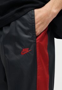 Κοντινό πλάνο χεριού στην τσέπη μαύρου και κόκκινου παντελονιού Nike, δείχνοντας το κόκκινο λογότυπο Nike σε μαύρο ύφασμα με κόκκινη πλαϊνή λωρίδα.