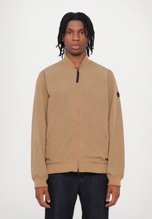Chaqueta bomber de color marrón claro, fabricada con tejido suave, que presenta un cierre de cremallera en la parte delantera, puños acanalados y un parche de logo redondo en la manga izquierda.