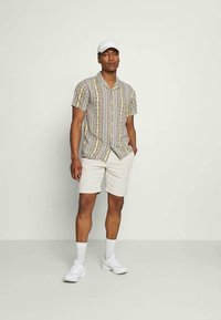 Uomo con berretto bianco, camicia a maniche corte con motivo, pantaloni corti beige, calze bianche e sneakers bianche, in piedi con le mani nelle tasche.