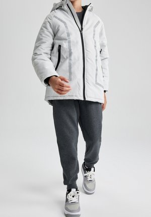 Personne portant une veste à capuche blanche avec des bandes réfléchissantes, un pantalon gris foncé et des baskets grises et blanches marchant vers l'avant.
