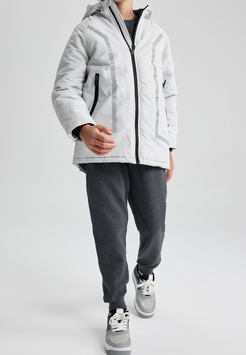 Personne portant une veste à capuche blanche avec des bandes réfléchissantes, un pantalon gris foncé et des baskets grises et blanches marchant vers l'avant.