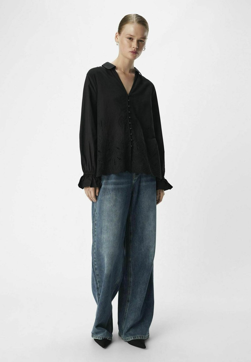 Une femme se tient debout portant un blouse noire à manches longues et à boutons, ainsi que des jeans larges bleus sur des chaussures noires à talons pointus.