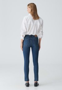 Jeans skinny en denim bleu foncé, avec une taille mi-haute, cinq poches et une patch en cuir marron à l'arrière. Associés à une chemise blanche.