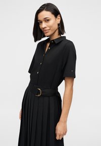 Μαύρο φόρεμα-πουκάμισο με κοντά μανίκια, χρυσά κουμπιά και ζώνη στη μέση με στρογγυλή αγκράφα. Η υφασμάτινη πλισέ φούστα προσθέτει λεπτομέρεια.