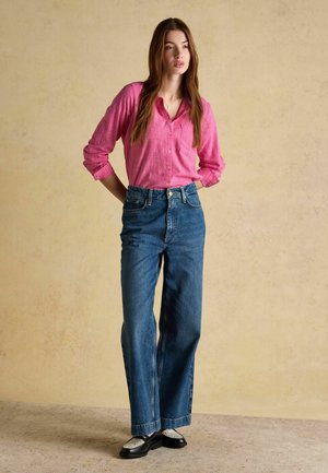 Sieviete stāv pret bēšas krāsas sienu, valkājot rozā pogājamu krekliņu, augstajiem jostasvietas zilajiem džinsiem un melnām loafer tipa kurpēm ar baltām zeķēm, rokas aiz muguras.