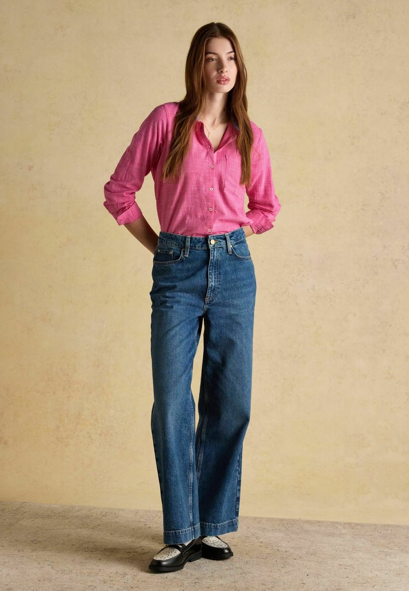 Mujer de pie contra una pared beige, lleva una camisa rosa con botones, vaqueros azules de cintura alta y mocasines negros con calcetines blancos, con las manos detrás de la espalda.