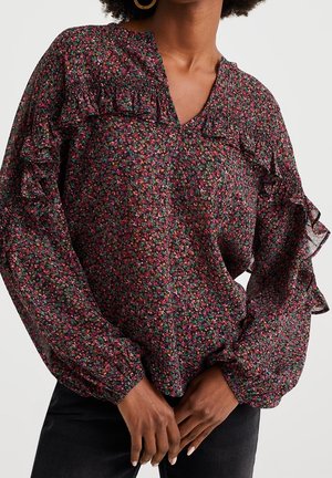 Vrouw draagt een donker bloemenblouse met lange mouwen en ruches op schouders en mouwen, gecombineerd met een zwarte broek en gouden creolen.