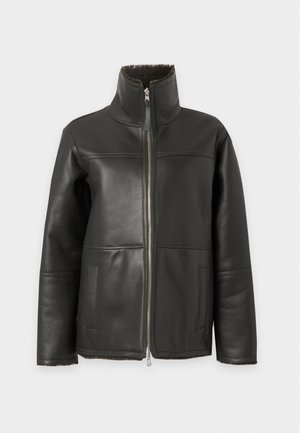 Γυναίκα που φοράει μαύρο σακάκι από shearling, πράσινο καρό πουκάμισο, φαρδιά σκούρα μπλε παντελόνια και μαύρα μοκασίνια, στέκεται μπροστά σε απλό φόντο.